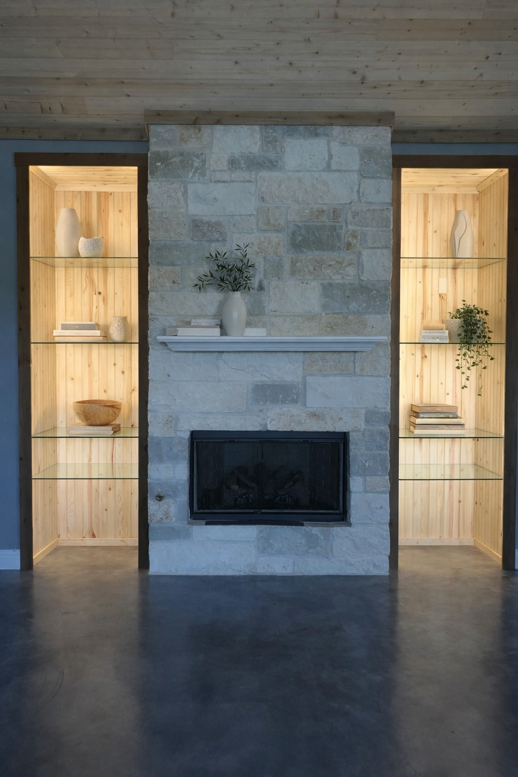 Living room fireplace with a stone surround, flanked by built-in wooden shelving units with glass shelves and decorative items inside, including vases, books, and a plant.