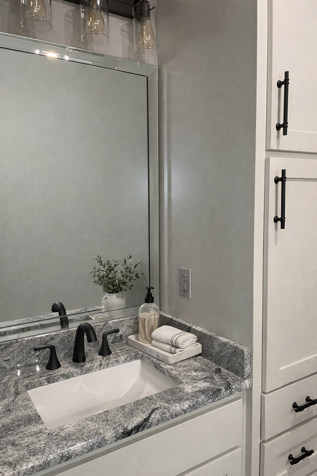 Bathroom vanity with granite countertop, black fixtures, potted plant, soap dispenser, folded towels, and large mirror.