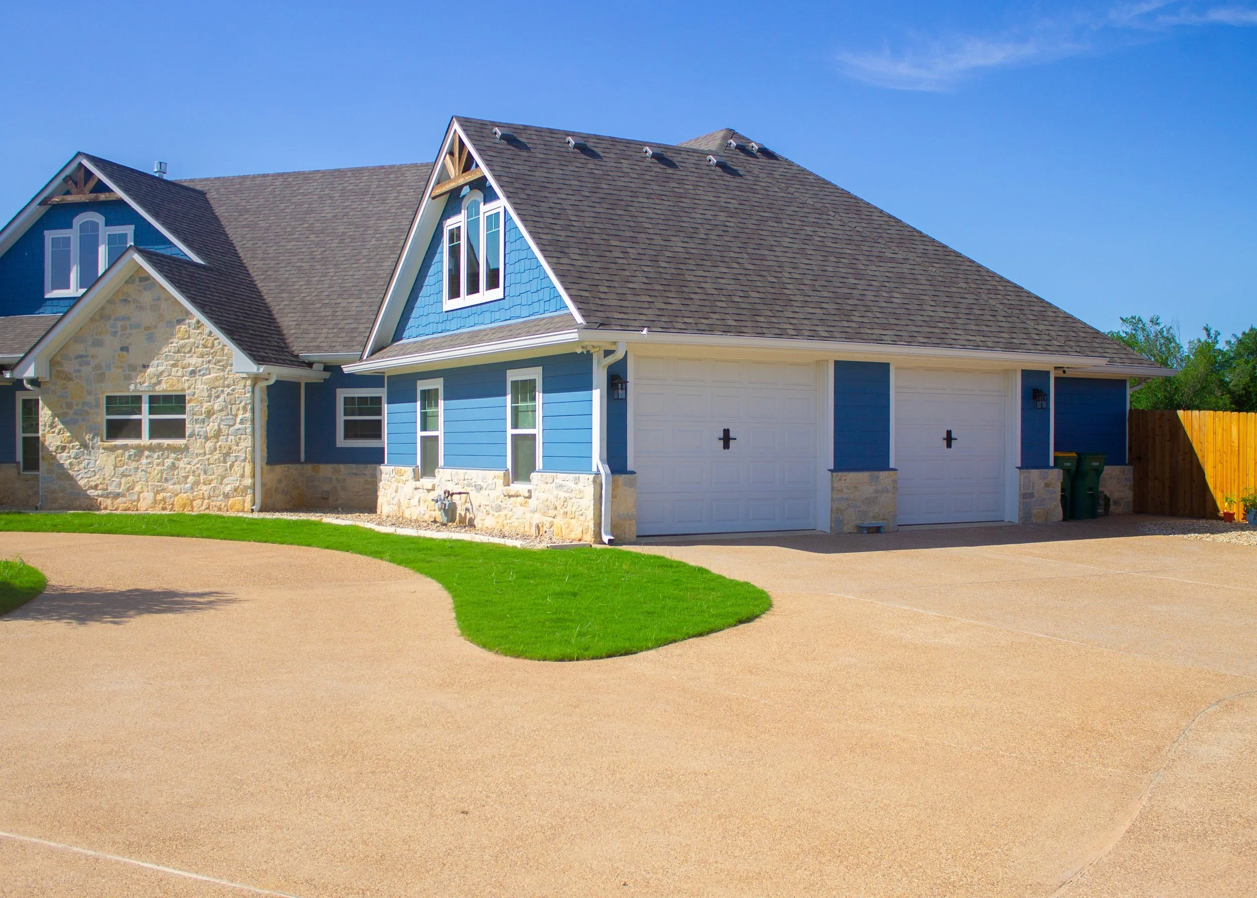 A detached house with blue and stone exterior, two garage doors, and a curved driveway surrounded by green grass and a wooden fence under a clear blue sky.