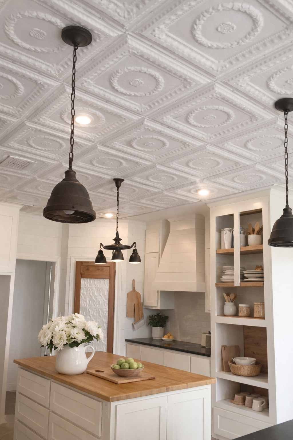 A kitchen with a white island topped with a vase of white flowers and a bowl of green apples. The ceiling has decorative white tiles and hanging black pendant lights. There are white cabinets with open shelves holding dishes and kitchen utensils.