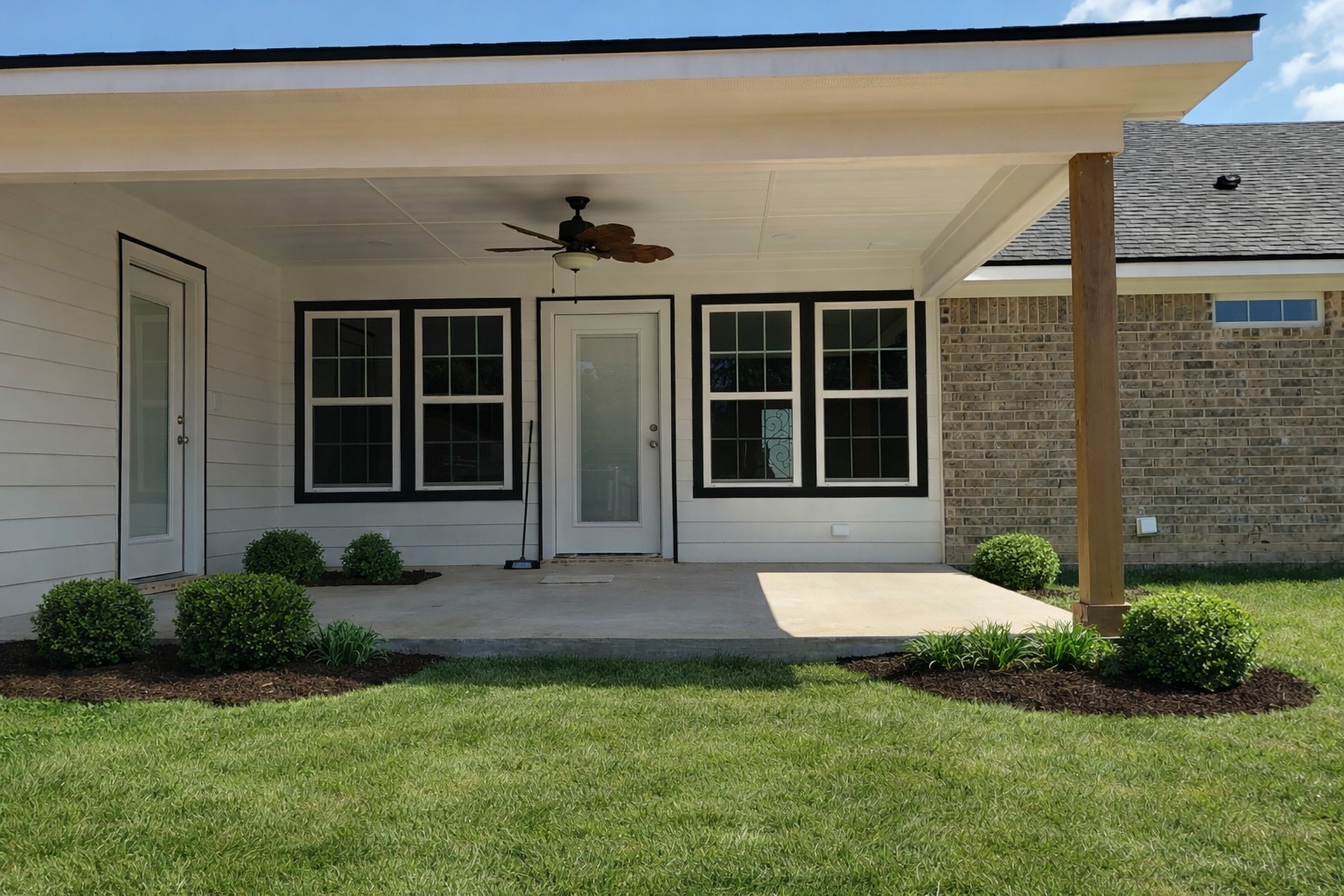 Back porch of a house with white siding, black window frames, and a glass door. There is a ceiling fan with leaf-shaped blades, a broom leaning against the wall, and landscaped bushes with green grass in the foreground.
