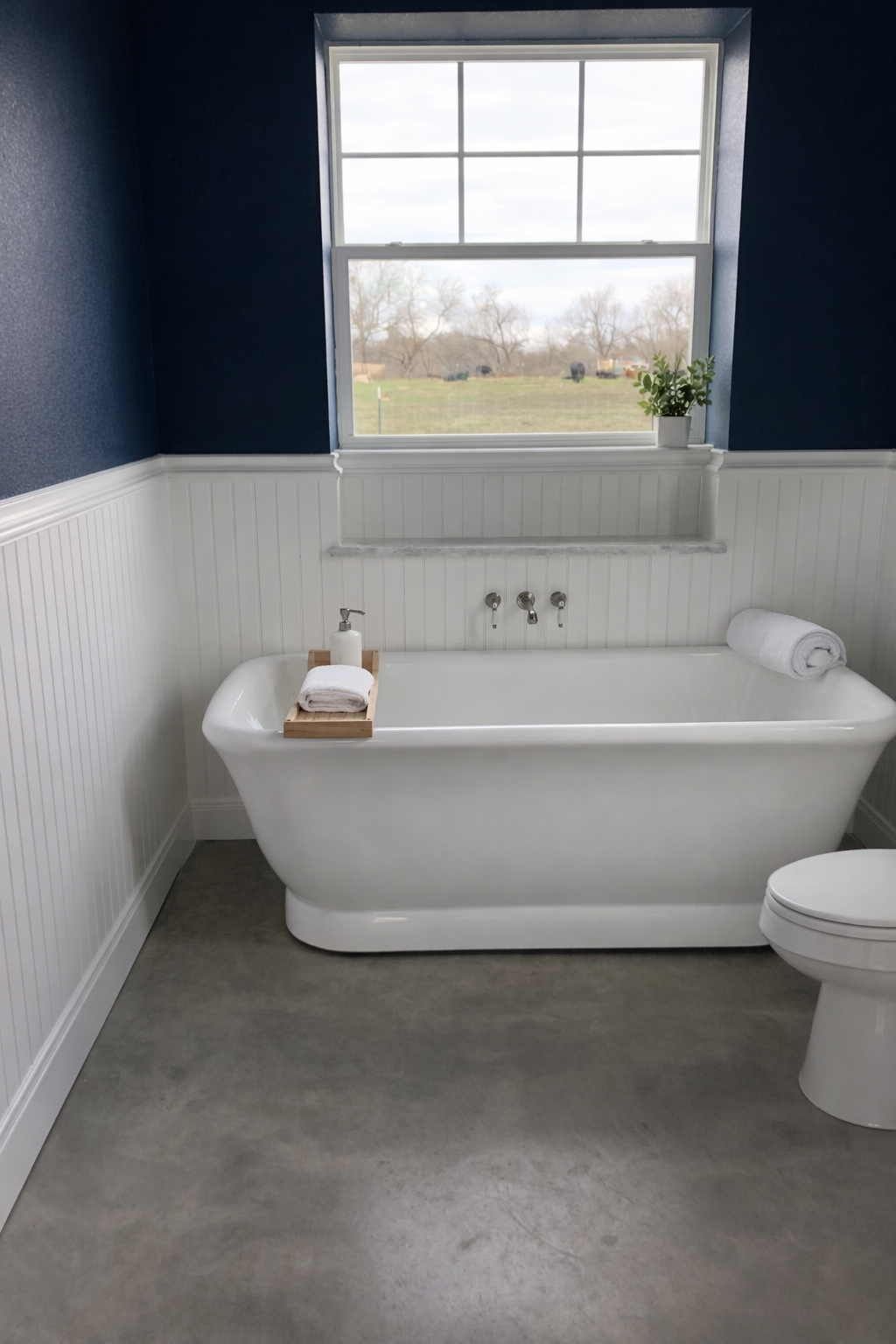 A bathroom with a white clawfoot bathtub below a large window, a small potted plant on the window ledge, and a rolled towel on the tub, with a view of a field outside.