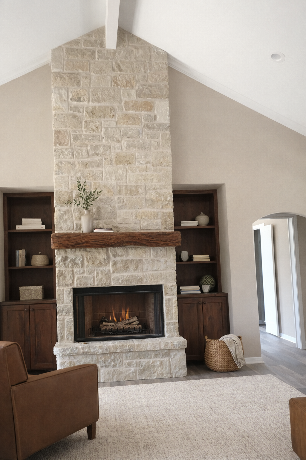 Living room with a stone fireplace, wooden mantel, built-in wooden bookshelves on each side, and a brown armchair in the foreground. The fireplace has a fire burning, and there are decorative items on the shelves and mantel, including a vase with gre