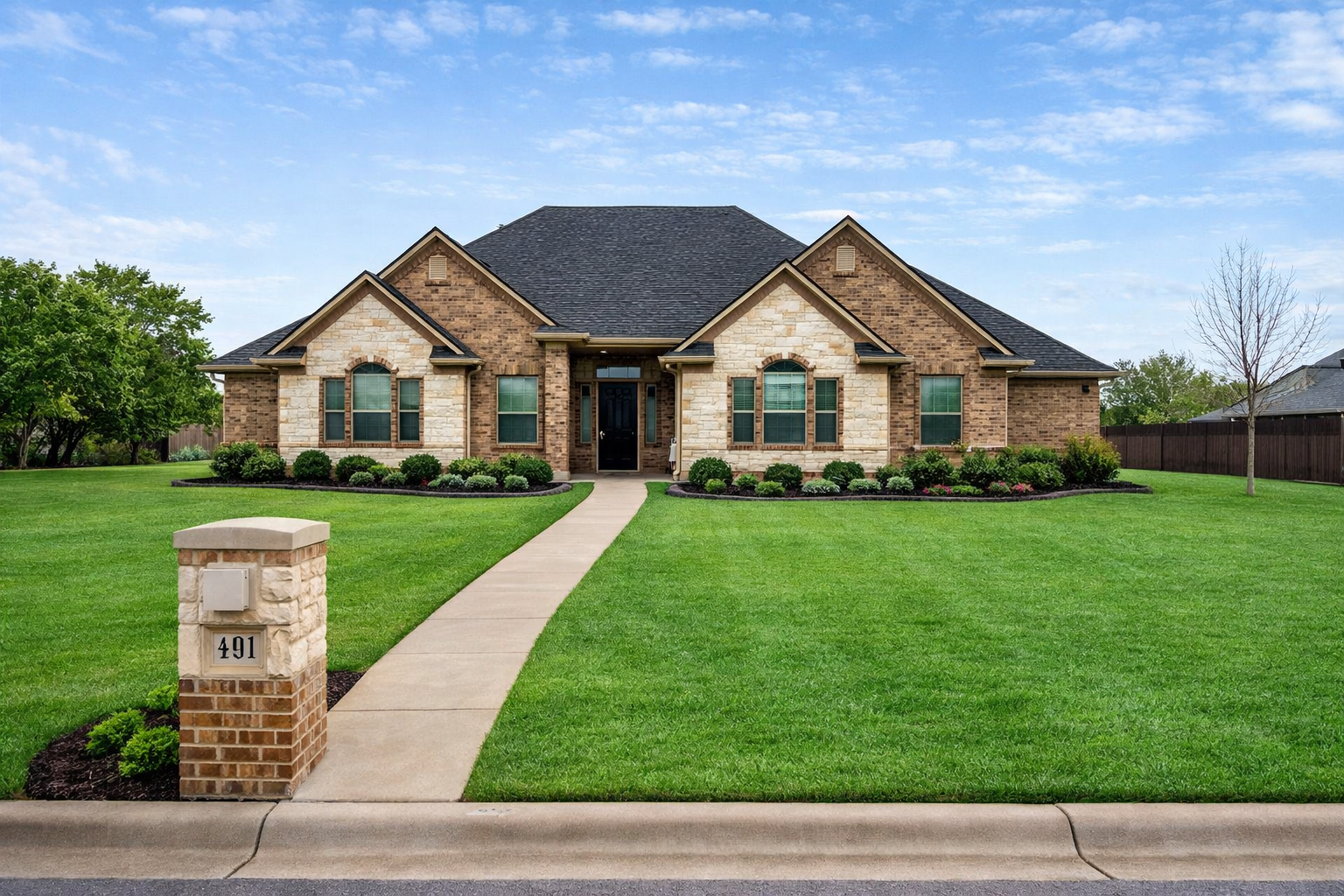 Front view of a suburban house with brick facade and well-maintained lawn, sidewalk leading to front door, house number 491, trees and a clear sky.