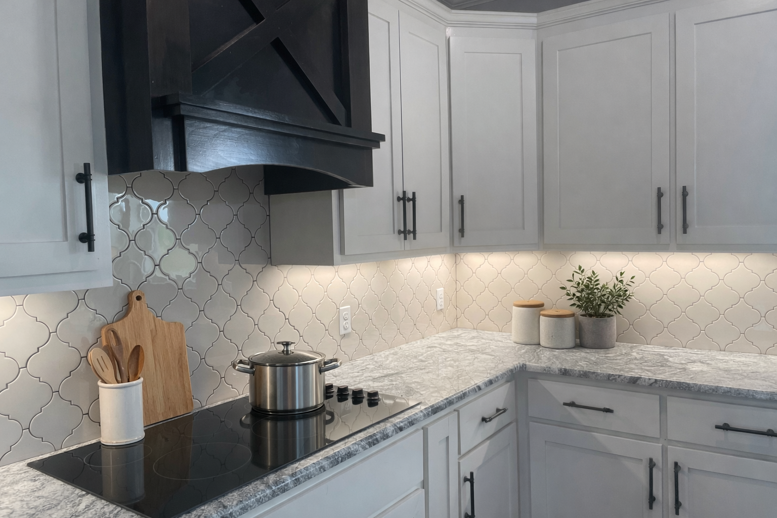 Modern kitchen with light gray cabinets, a black range hood, a white and gray patterned backsplash, a marble countertop, a stainless steel pot on an electric stove, a wooden cutting board, and three small potted plants on the counter.