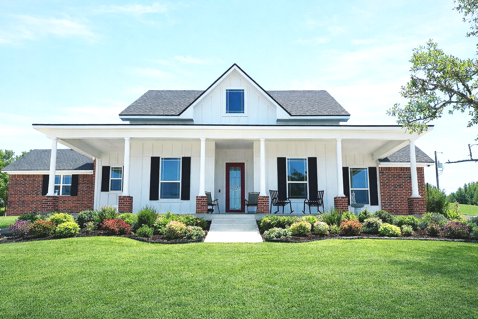 A two-story house with a front porch, white exterior with brick accents, black shutters, and a red front door, surrounded by a manicured lawn and colorful garden in bright sunlight.