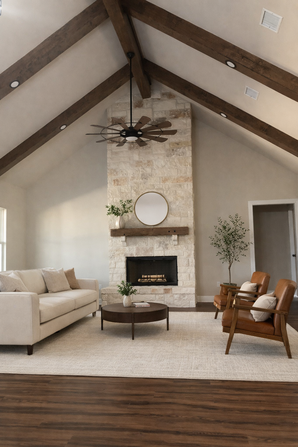 Living room with beige sofa, two brown armchairs, a round coffee table, and a white rug. Features a stone fireplace with a round mirror, potted plants, and a wooden mantel. Exposed wooden beams and ceiling fan.