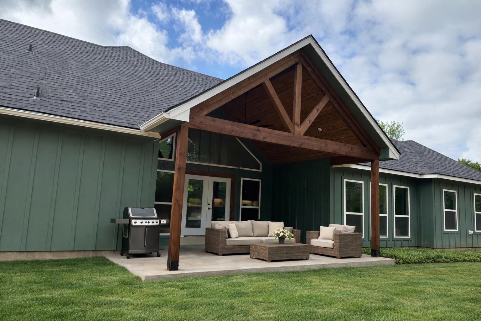 Backyard patio with outdoor furniture and grill, attached to a green house with large windows and a wooden gabled roof.