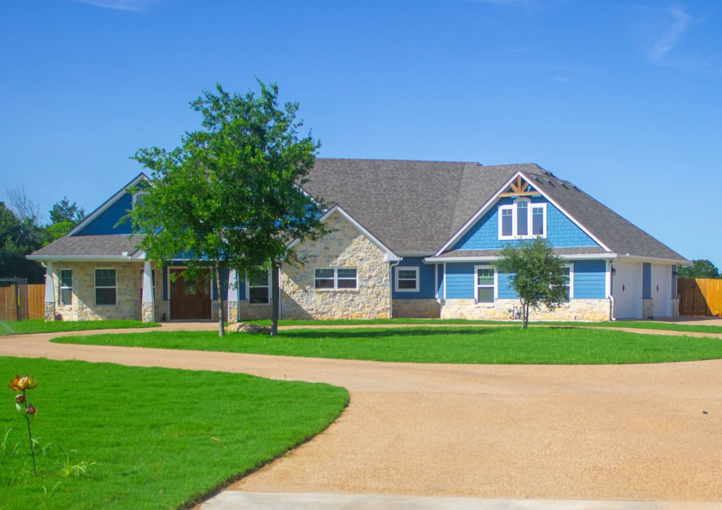 A large house with stone and blue siding, multiple windows, a brown front door, a spacious driveway, green lawn, and trees, under a clear blue sky.
