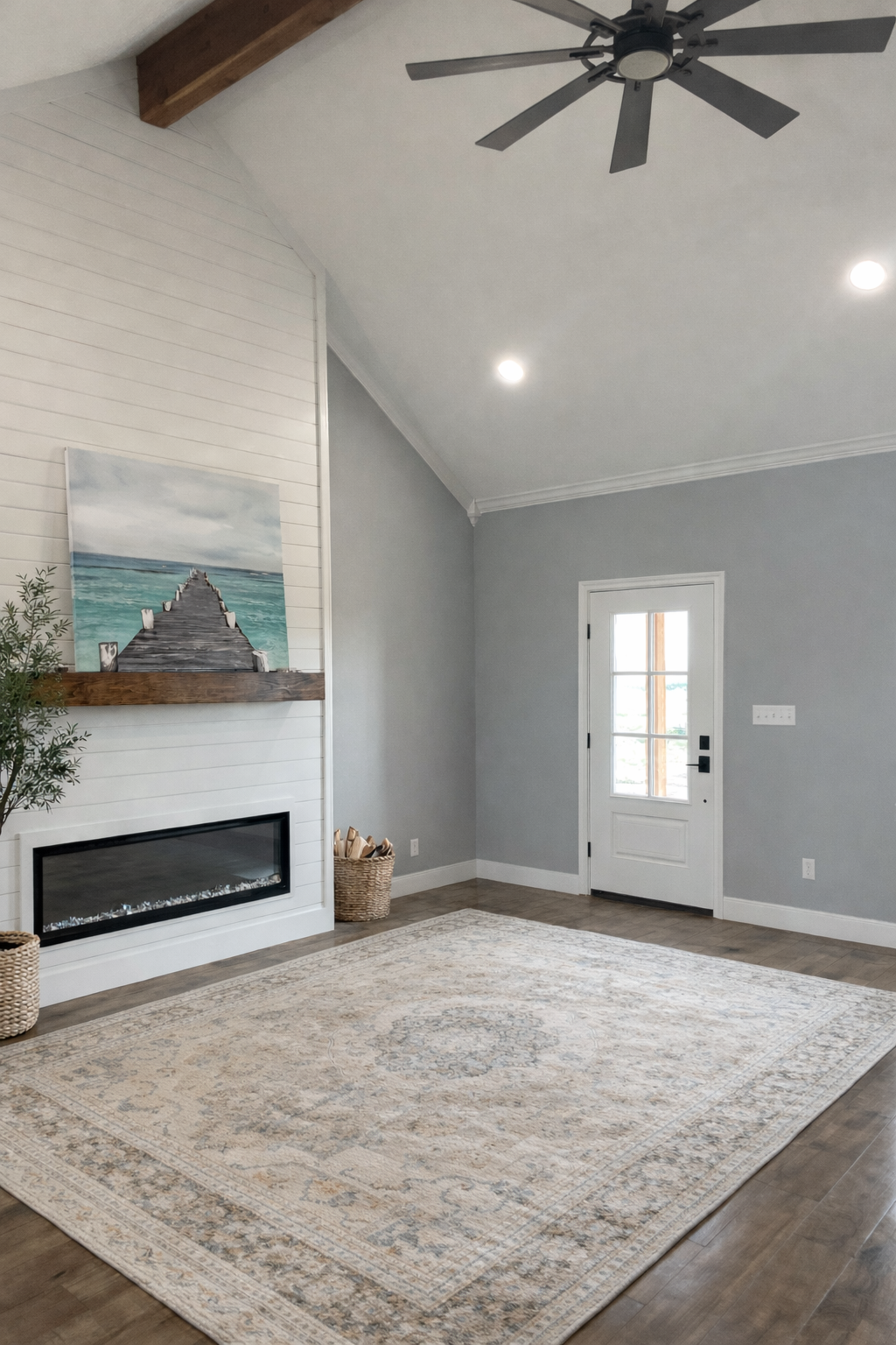 Living room with a white shiplap fireplace, a colorful painting of a dock extending into water, a woven basket with firewood, a light-colored patterned rug, a white door with glass panes, and a ceiling fan.