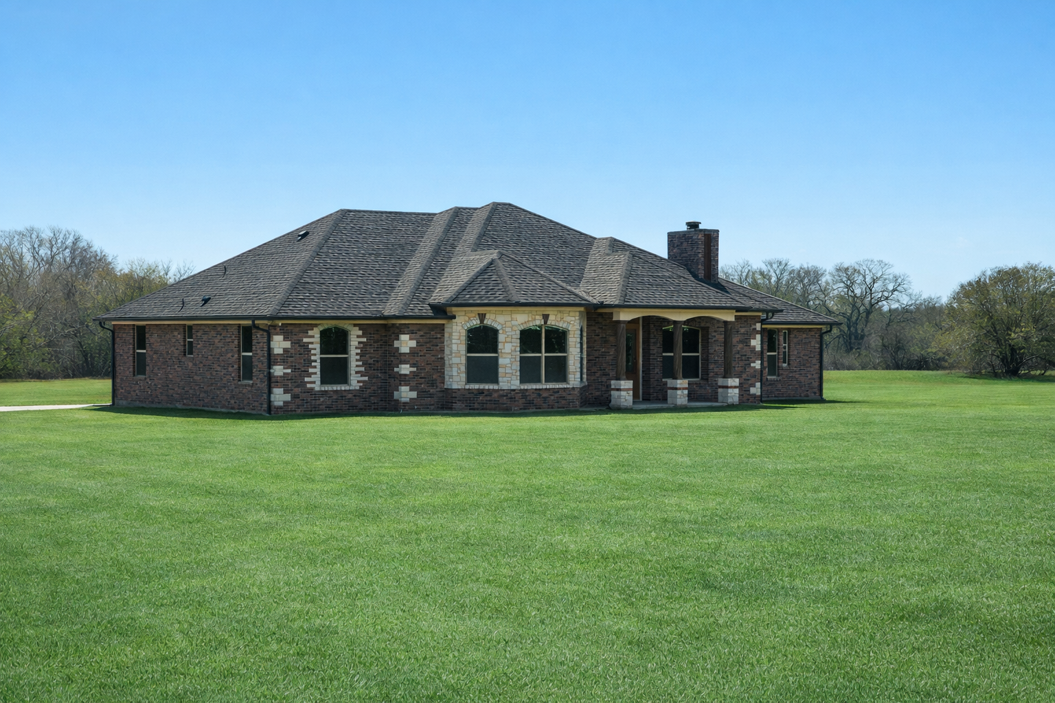 A large brick house with a dark shingle roof on a spacious, well-manicured lawn under a clear blue sky.