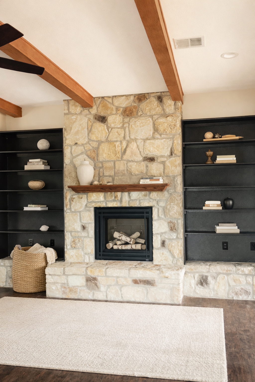 Living room with stone fireplace, black bookshelves on either side, decorative items, a beige rug, and a woven basket.