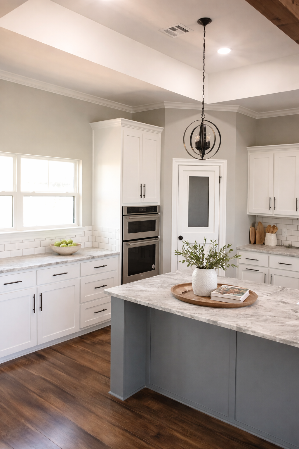 Modern kitchen with white cabinets and marble countertops, a center island with a plant and book, stainless steel oven, white subway tile backsplash, wooden flooring, and a black pendant light