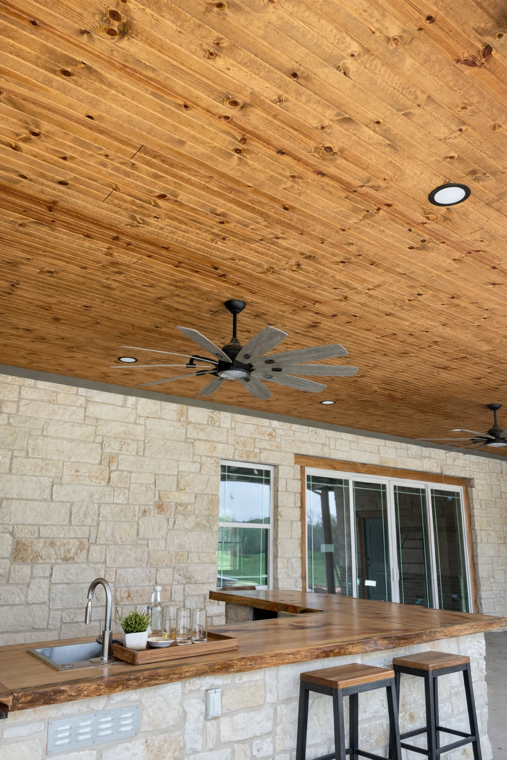 A kitchen with a stone wall, large windows, a wooden countertop, and two bar stools. The ceiling is wood-paneled with pendant lights and ceiling fans.