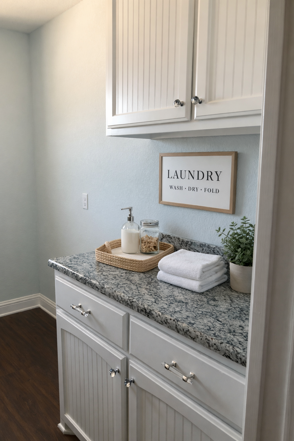 A laundry room with white cabinets, a granite countertop, a framed sign reading 'LAUNDRY Wash, Dry, Fold,' a soap dispenser, a jar of cookies, folded white towels, and a potted plant.