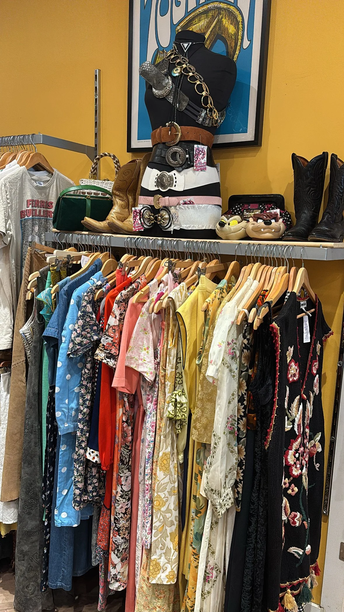 Clothing and accessories displayed in a vintage or thrift store, including dresses, belts, boots, and jewelry, with a colorful poster in the background.