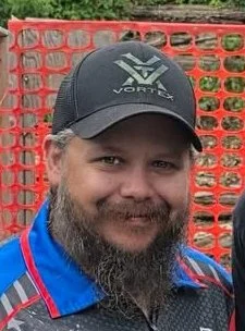 A man with a beard wearing a black cap and a blue jacket, standing in front of an orange construction fence.