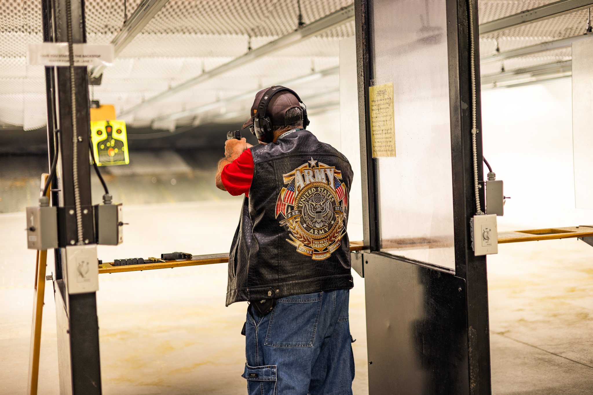 A man wearing a leather jacket with an Army logo on the back and a black baseball cap is at an indoor shooting range, aiming a handgun at a target downrange.