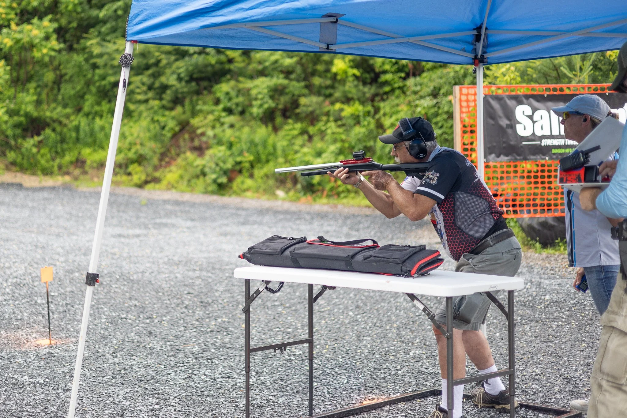 A man aiming a rifle at a shooting range under a blue canopy with another person nearby, and trees in the background.