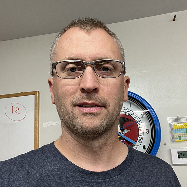 A man with glasses and short hair taking a selfie indoors, with a whiteboard, a clock, and a calendar on the wall behind him.