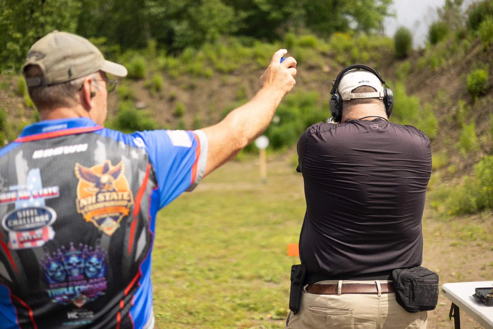 Two men practicing target shooting outdoors with guns, one pointing a gun and the other aiming, against a backdrop of green trees and grass.