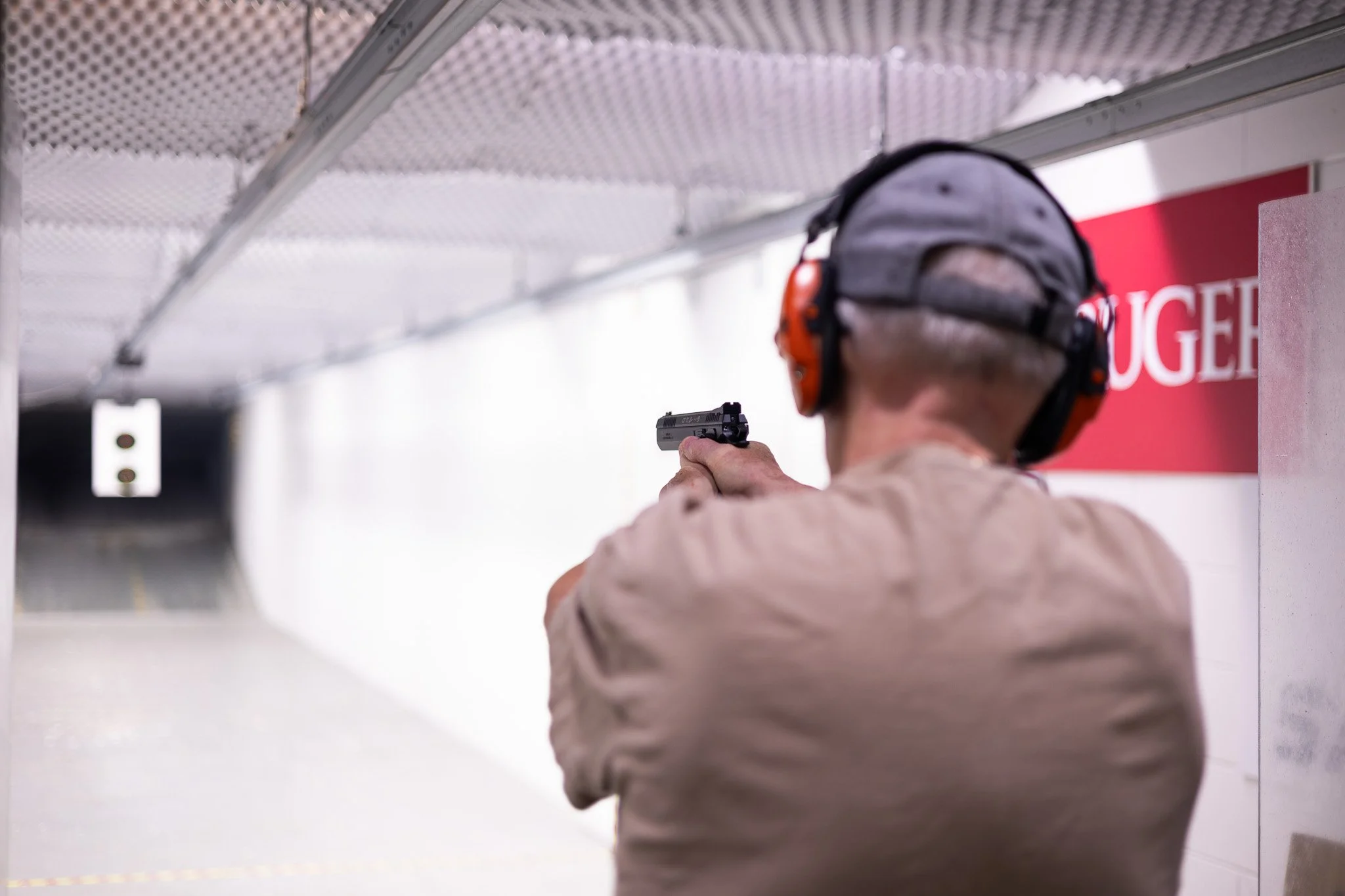 A person at an indoor shooting range aiming a handgun while wearing ear protection and a cap.
