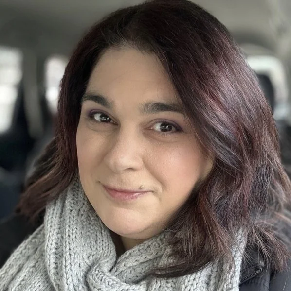 A woman with shoulder-length wavy hair and a light scarf inside a vehicle.