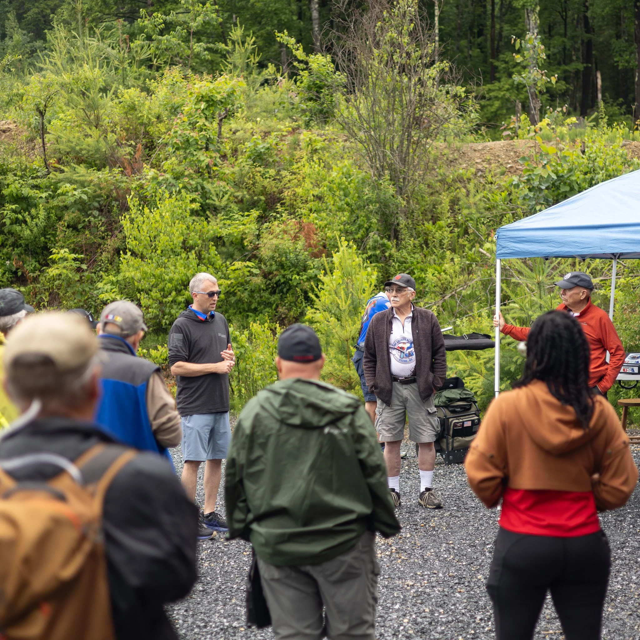 A group of people gathered outdoors under a green, forested area, some wearing caps and sunglasses, with one person holding a blue canopy tent. The scene appears to be a social or community event.