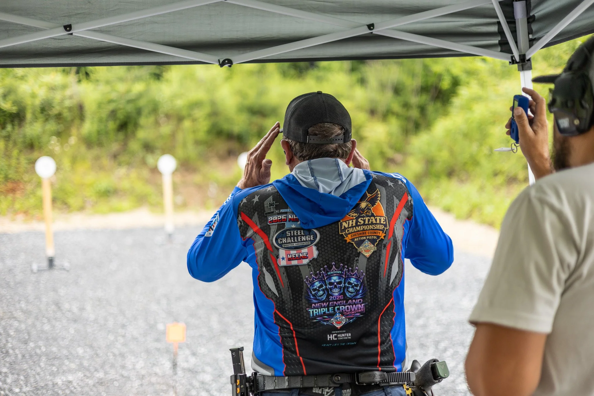 Two men at a shooting range, one adjusting his hat and the other holding a semi-automatic pistol. The back of the man wearing a blue jacket with various patches and a hat and the other man wearing a helmet and holding a blue device. Target stands are visible in the background.
