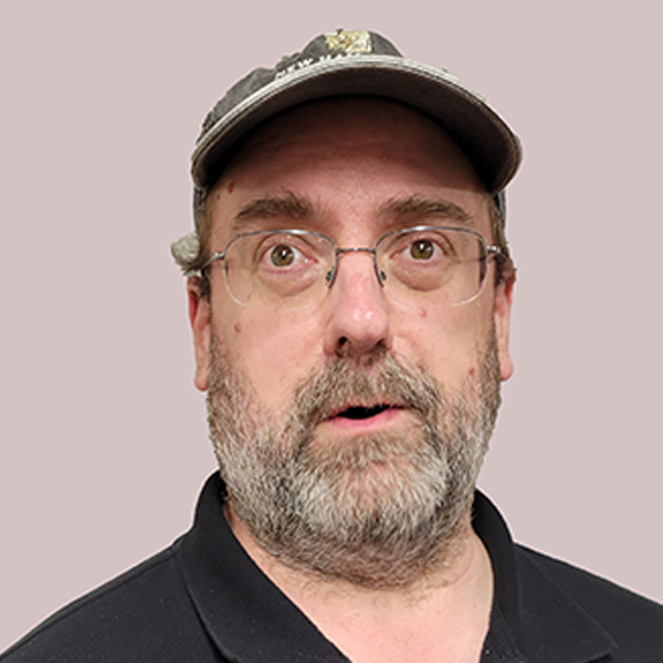 A middle-aged man with glasses and a beard, wearing a camouflage baseball cap and a black shirt, looking surprised or curious.