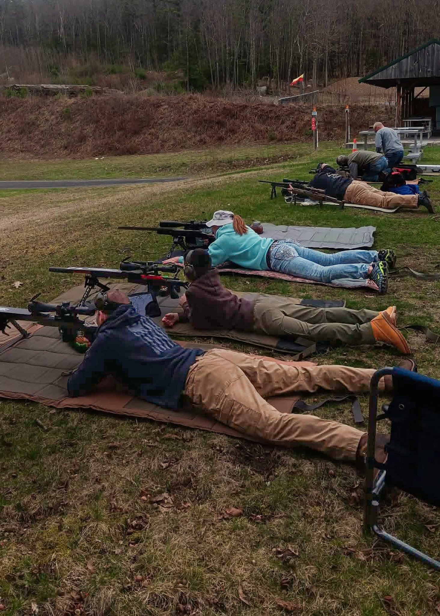 A person aiming a rifle during outdoor shooting practice, with another person guiding or recording, in a natural setting.