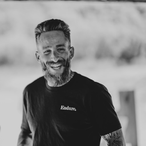 A smiling man with a beard and styled hair, wearing a black t-shirt, standing outdoors against a blurred background.