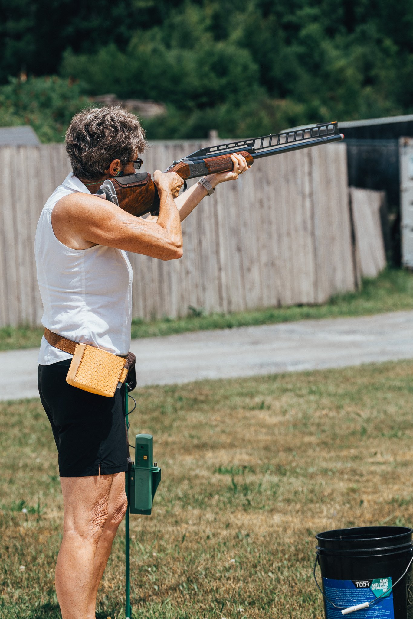 An older woman aiming a shotgun outdoors in a yard with a wooden fence and trees in the background.