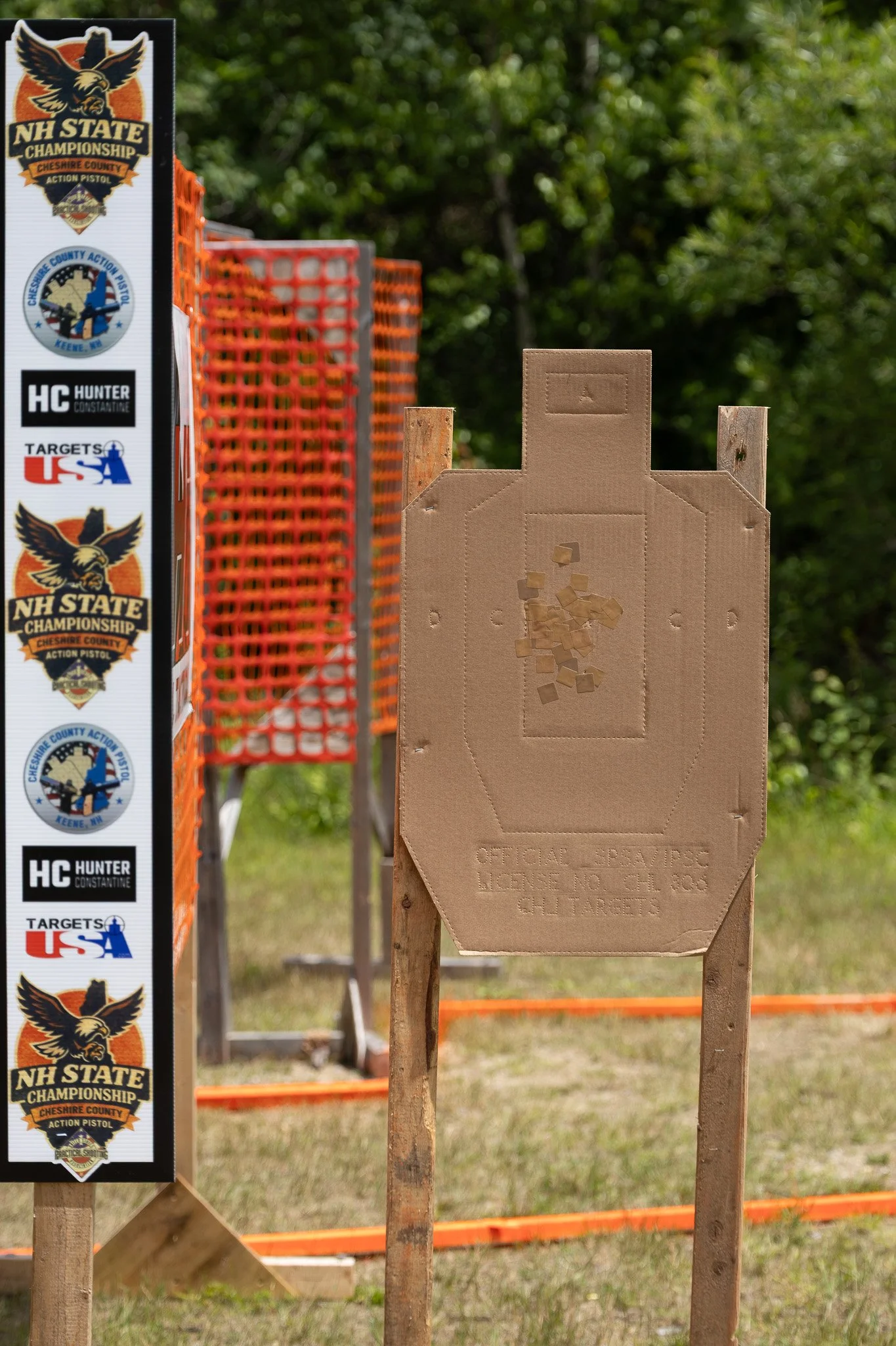 A cardboard target with a bullet hole in the upper center, set on wooden support at an outdoor shooting range. To the left, a vertical strip displays badges and stickers related to NH State Championship shooting events. In the background, orange safety barriers and green trees are visible.