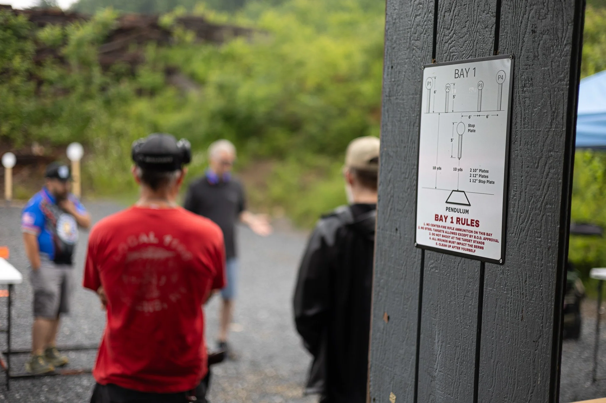 A group of people gather outdoors at a shooting range, with a sign on a dark wooden post providing rules for Bay 1, including target distances and safety instructions, surrounded by green trees in the background.