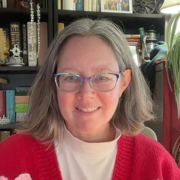 A woman with shoulder-length gray hair, wearing glasses, a white shirt, and a red sweater, smiling in front of a bookshelf filled with books and decorative items.