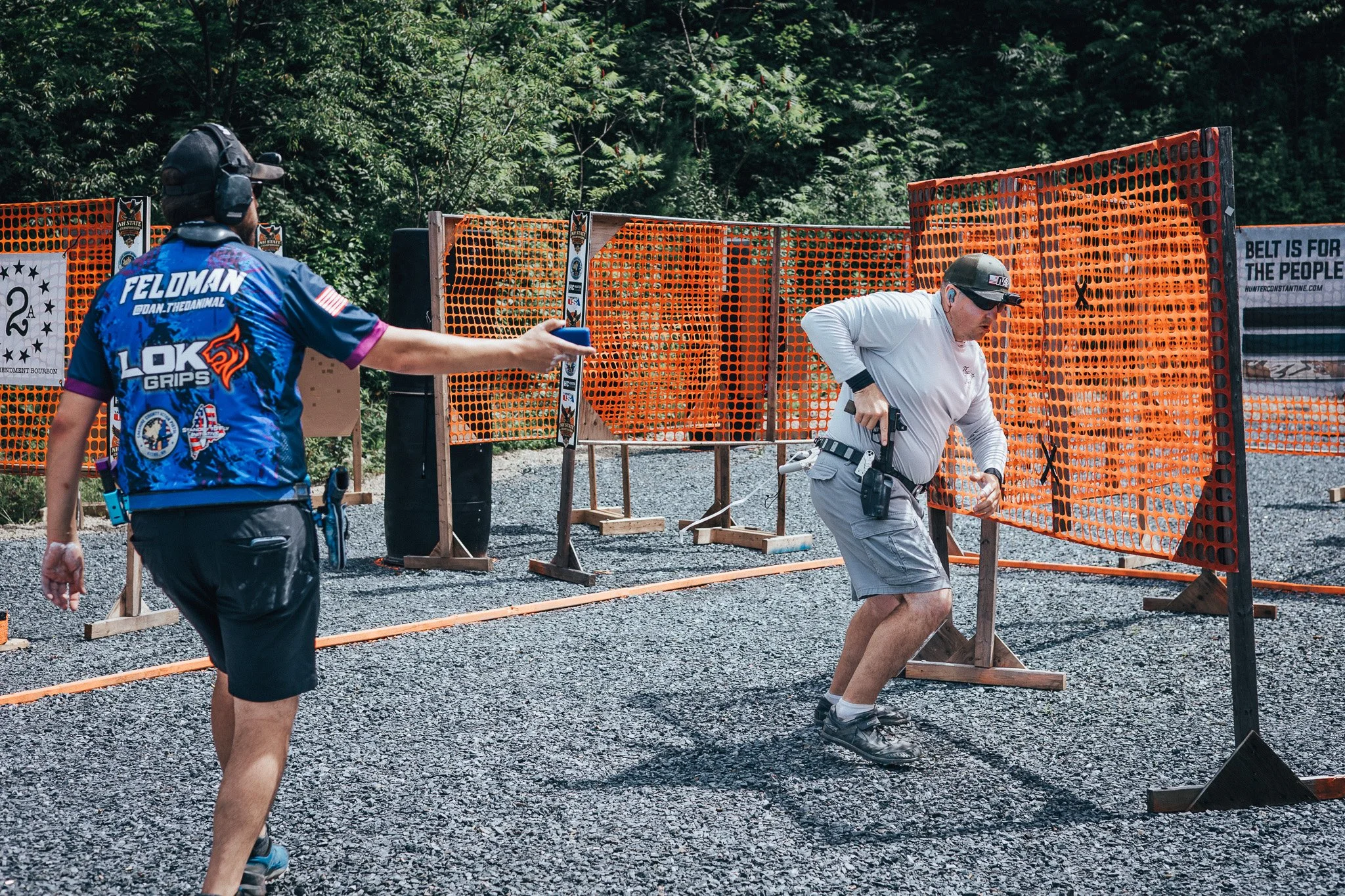 Two men are at a shooting range outdoors, with orange safety barricades and trees in the background. One man, wearing a blue shooting jersey with 'Feldman' on the back, is holding a smartphone, while the other man, dressed in white, appears to be adjusting his firearm.