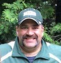 A man wearing a baseball cap and a green jacket, smiling outdoors with green trees in the background.