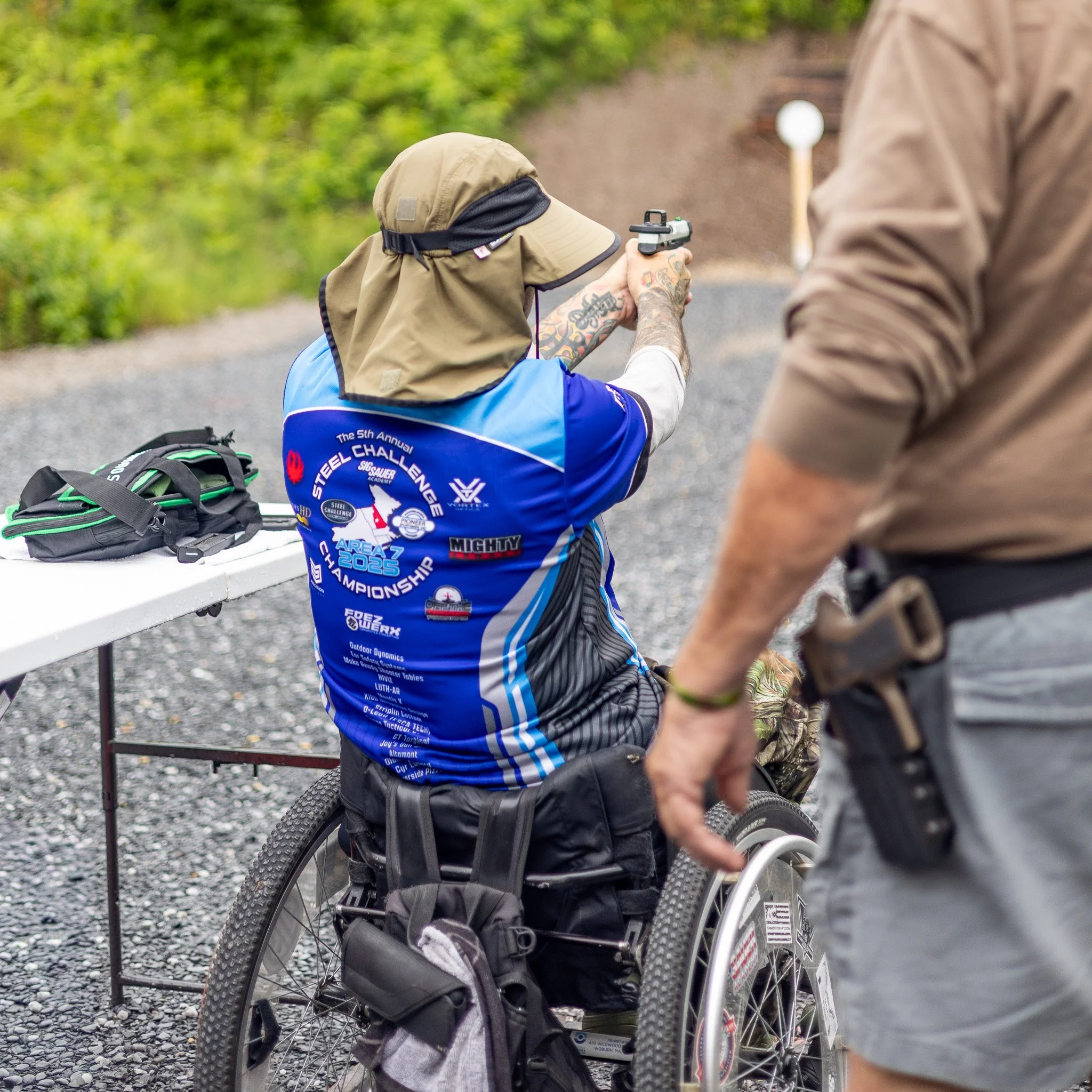 A person in a wheelchair aiming a handgun at a target outdoors. The person is wearing a baseball cap and a blue sports jersey. There is another individual nearby, partially visible. The background features greenery and a gravel surface.