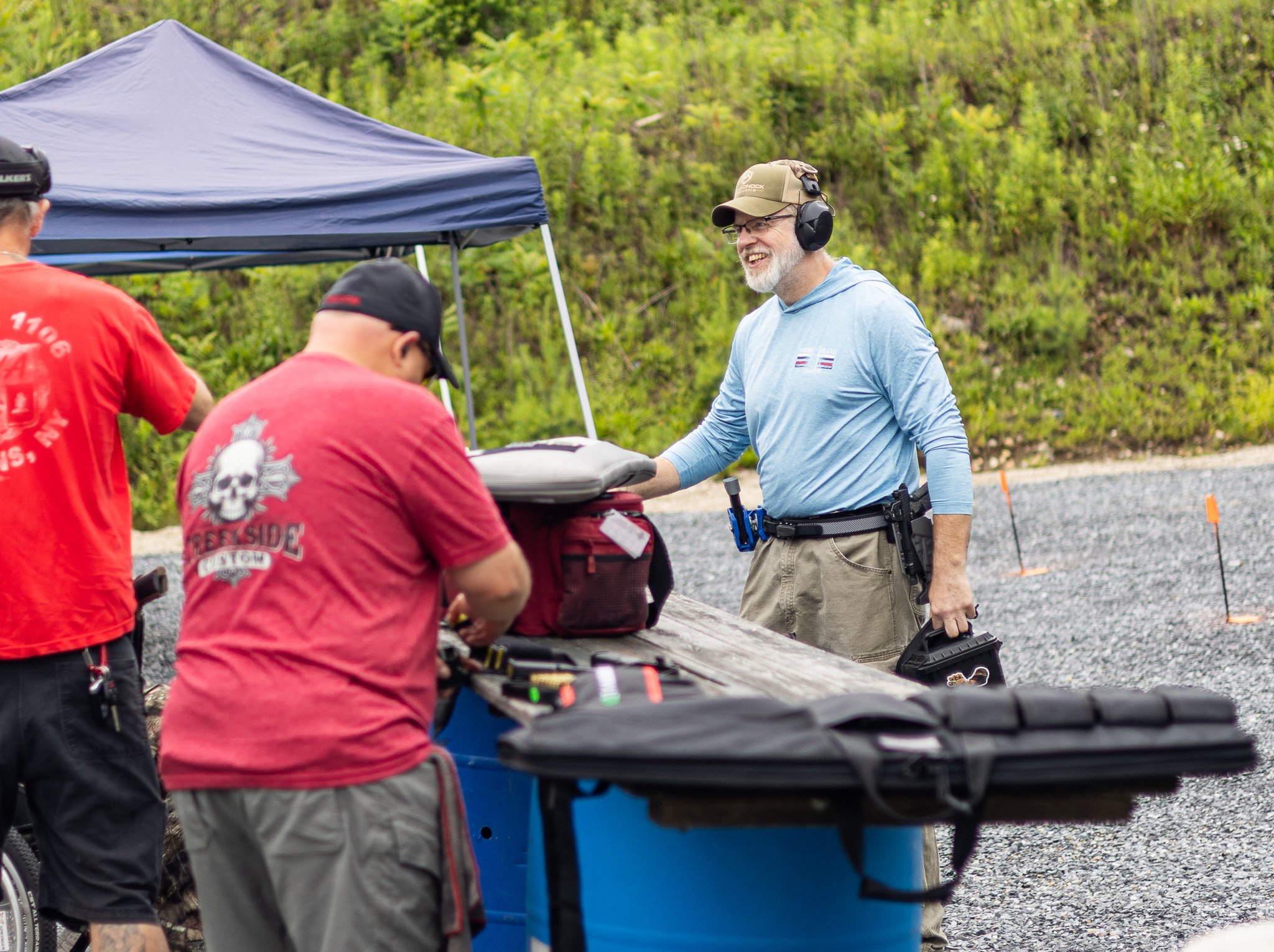 A group of firefighters working outdoors, with one man smiling and wearing hearing protection, standing near a blue portable fire pump or equipment, and other team members in red shirts and hats.