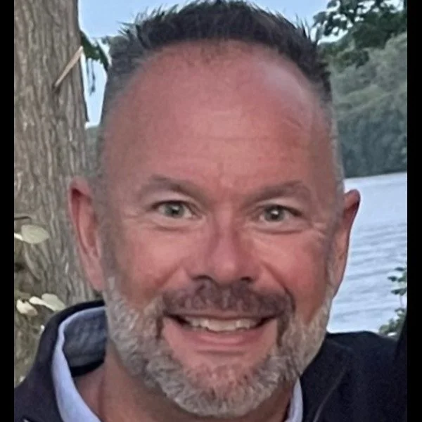 Close-up of a smiling man with short dark hair and a beard, standing outdoors near water and trees.