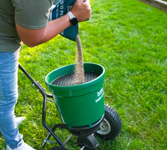 Person pouring granular fertilizer into a green yard cart on a grassy lawn.