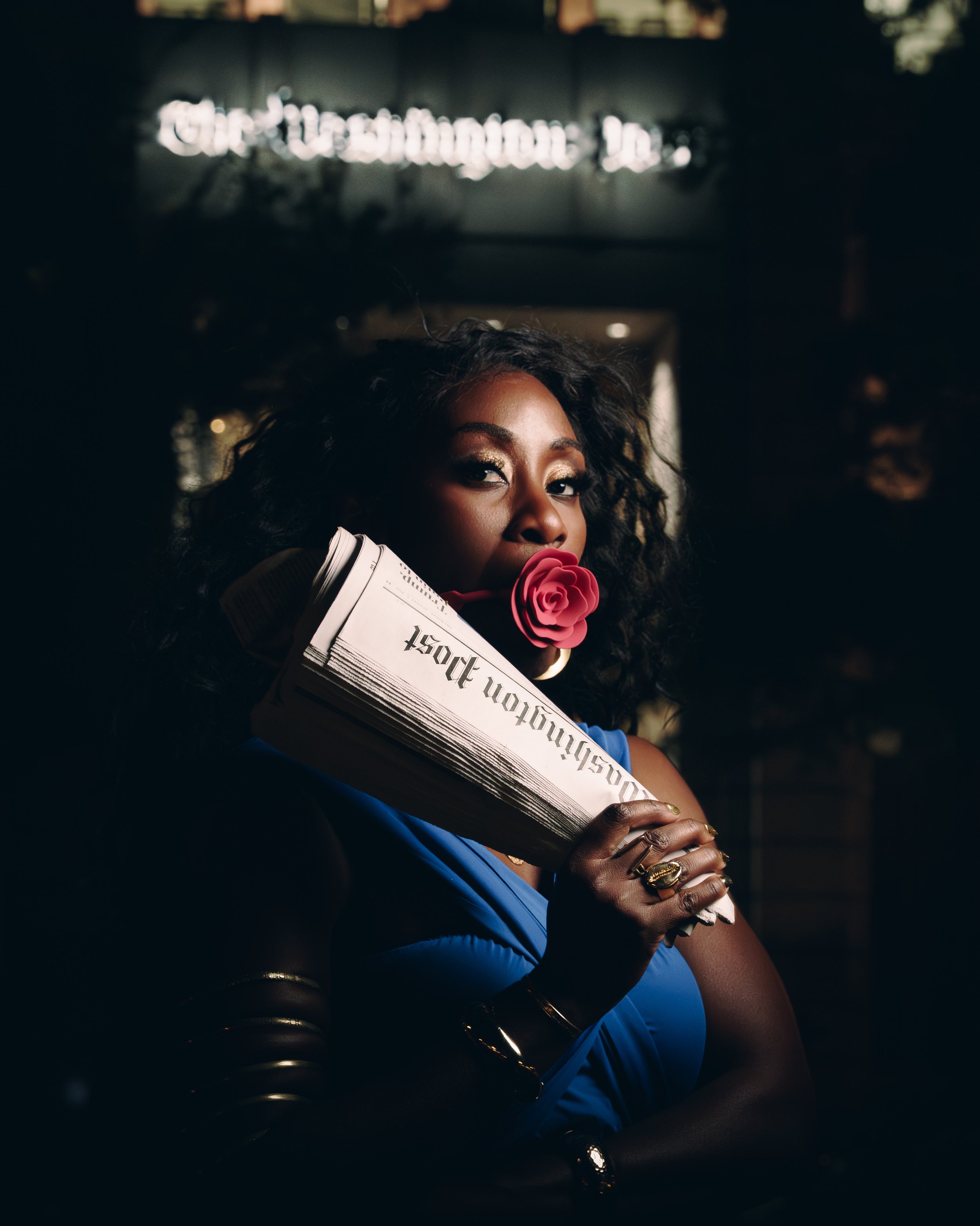 A woman with dark curly hair holding a pink rose in her mouth and newspapers in her hand, wearing a blue dress with gold jewelry, standing in front of a dark background with a sign that reads 'Washington'. 