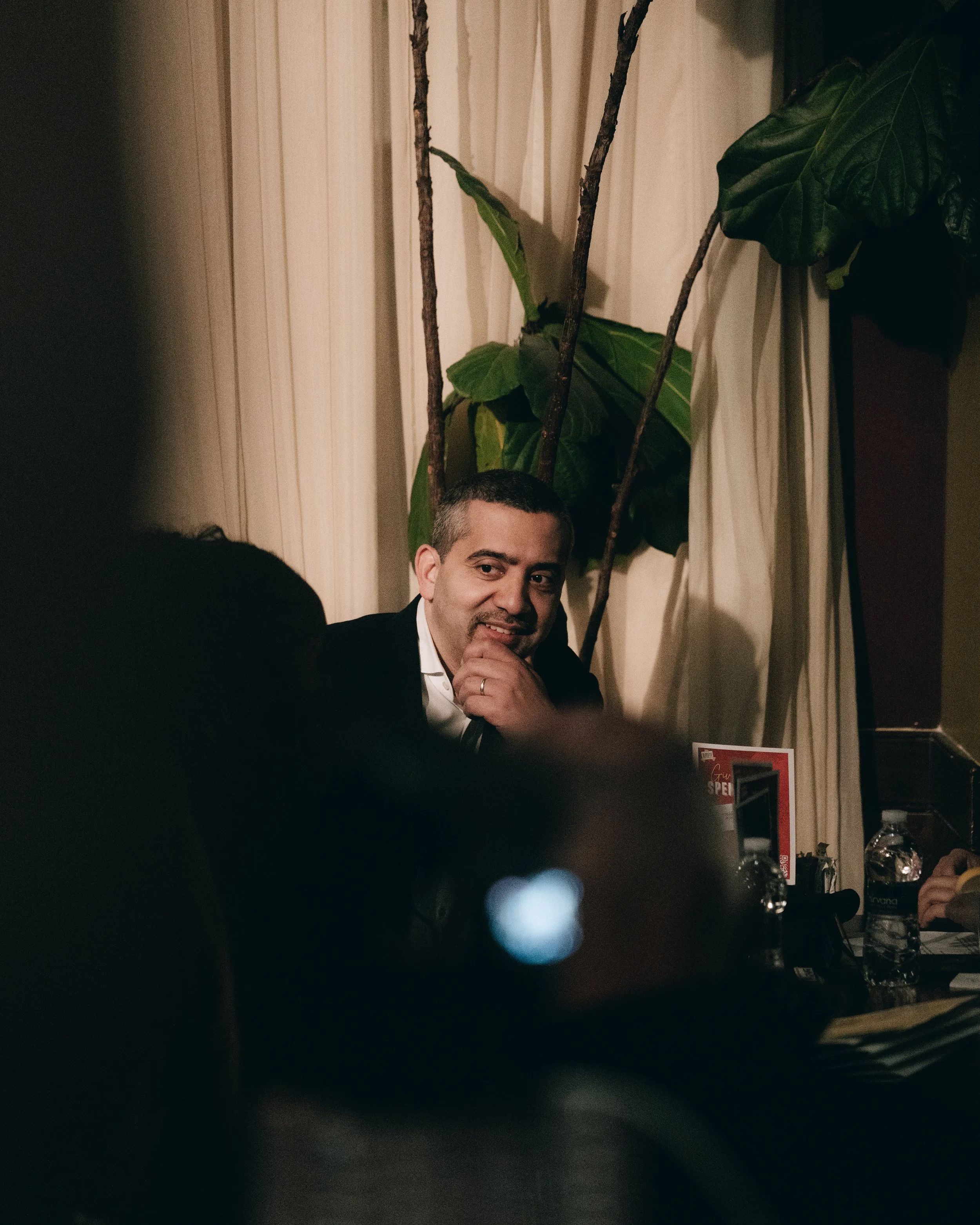 A man with short dark hair, wearing a dark suit and white shirt, sitting at a table with water bottles, a phone, and other items, leaning his chin on his hand, smiling, with large green plants behind him and curtains in the background.