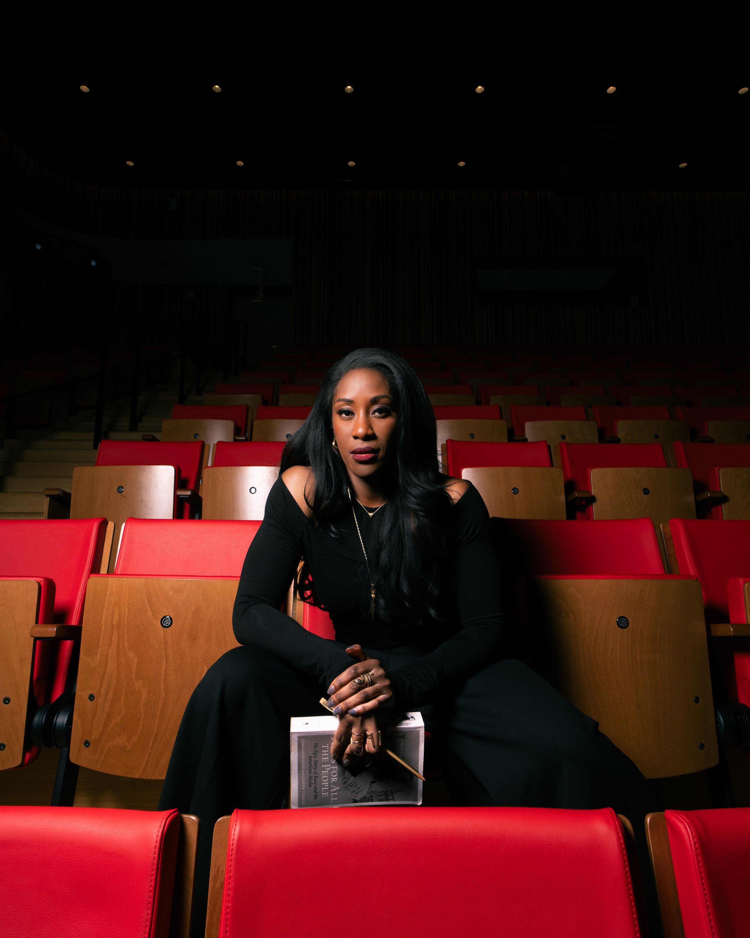 A woman with long black hair in a black off-shoulder dress sitting in an empty theater with red and wooden seats, holding a newspaper and a pencil.