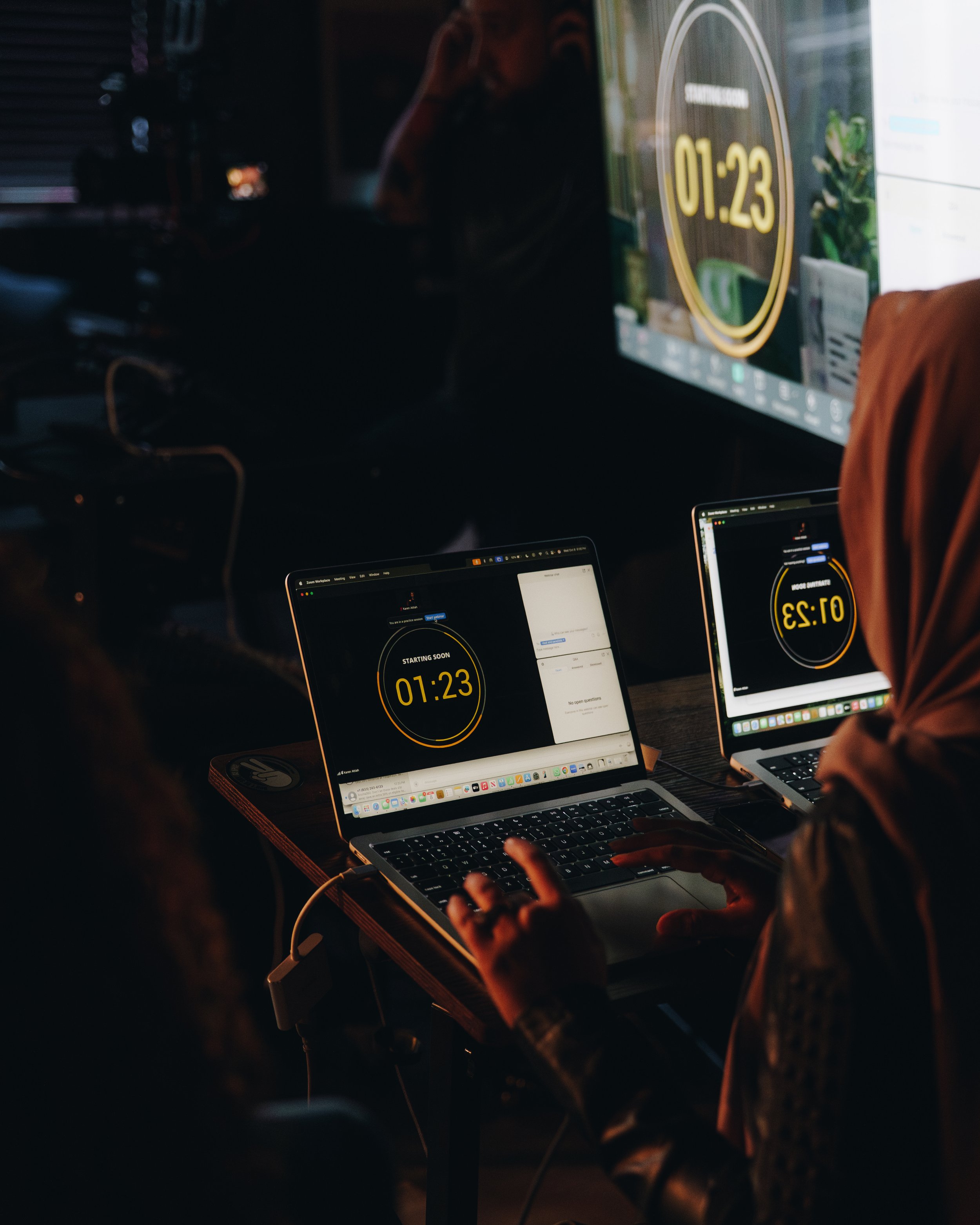 Two people working on laptops with visual countdown timers on their screens, in a dimly lit room.