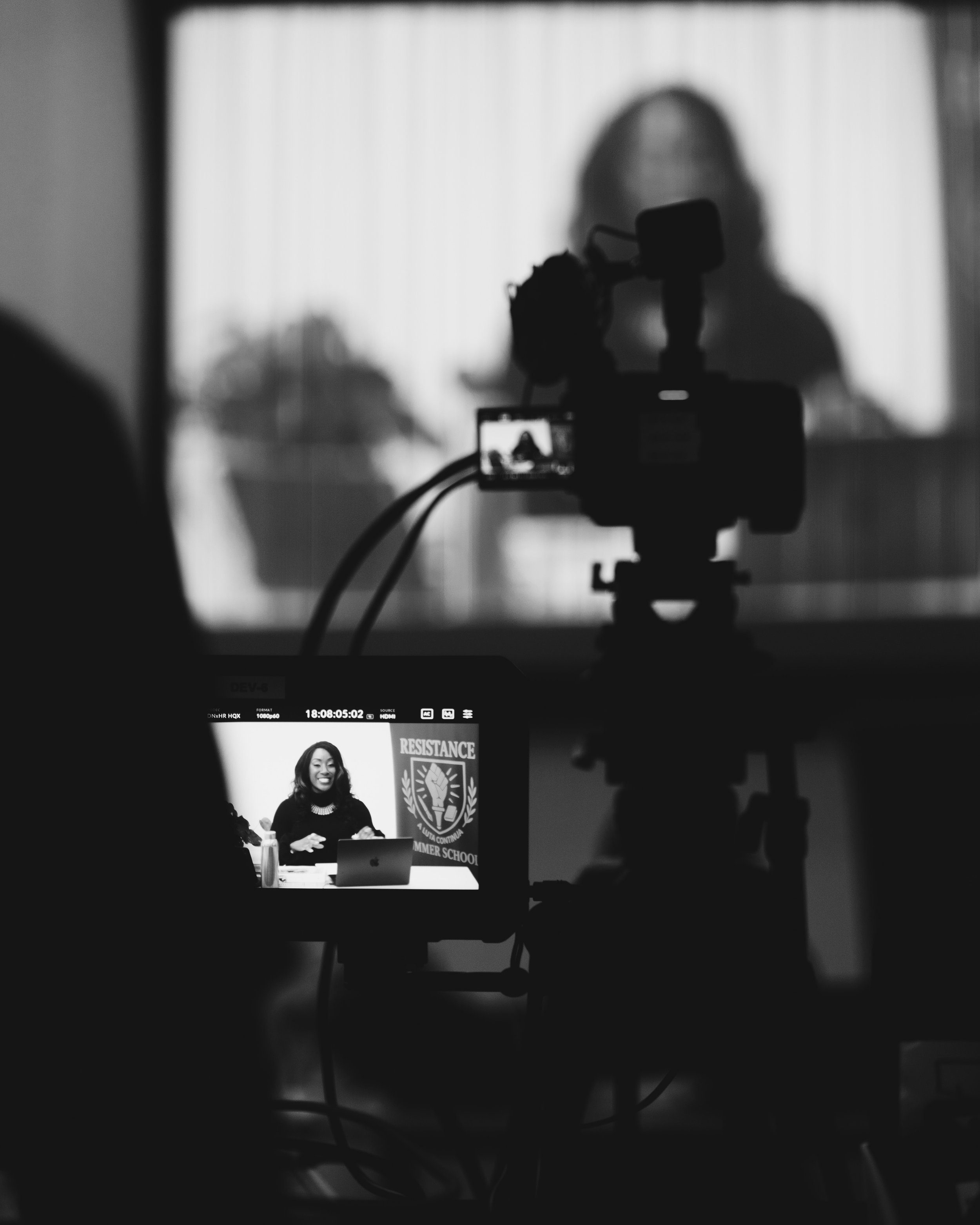 Black and white photo of a woman recording a video or live stream at a desk with a laptop, with a backdrop displaying a 'RESISTANCE' emblem.
