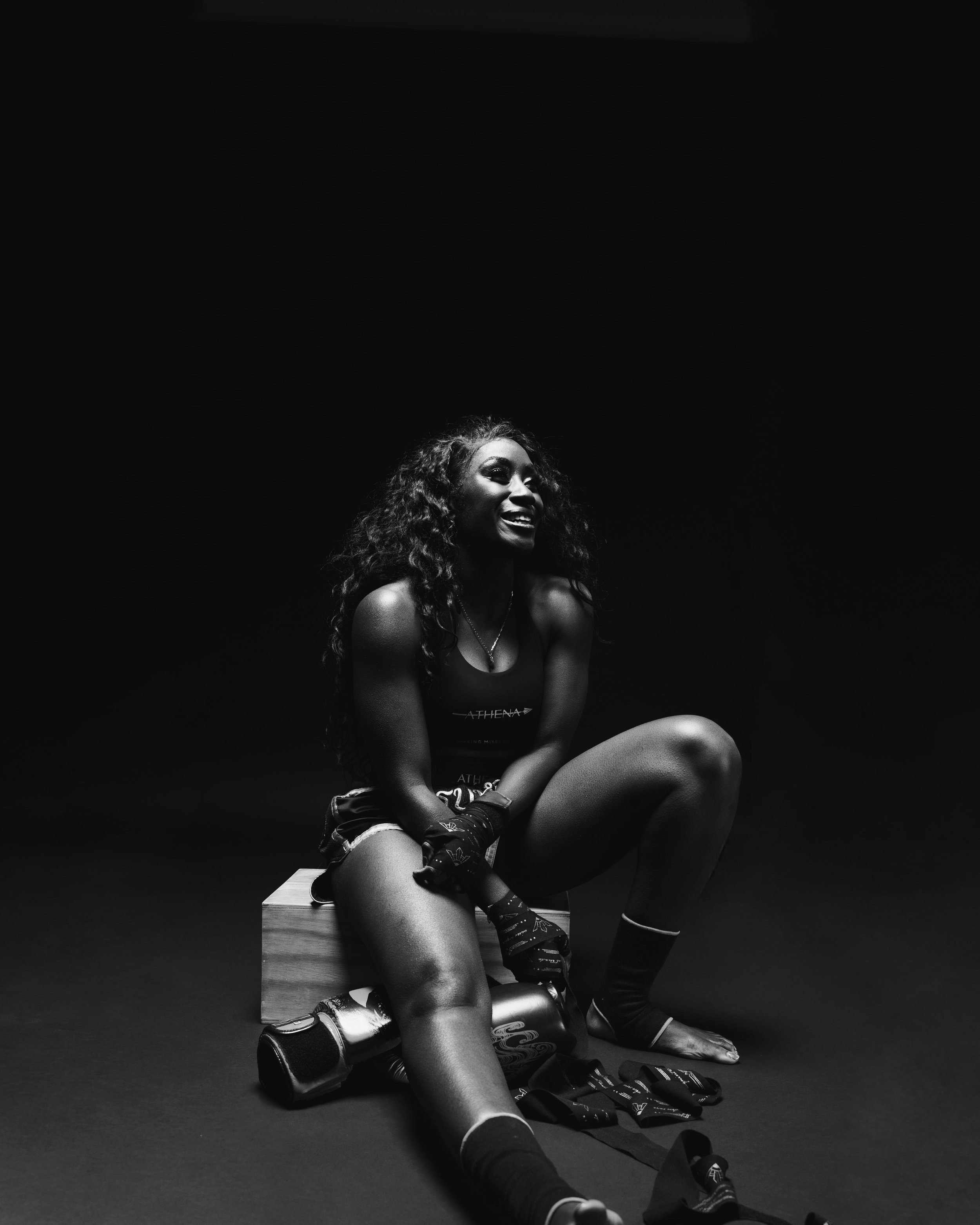 A woman sitting on a wooden box, dressed in athletic gear, smiling and surrounded by boxing gloves and wraps, in a dark studio setting.