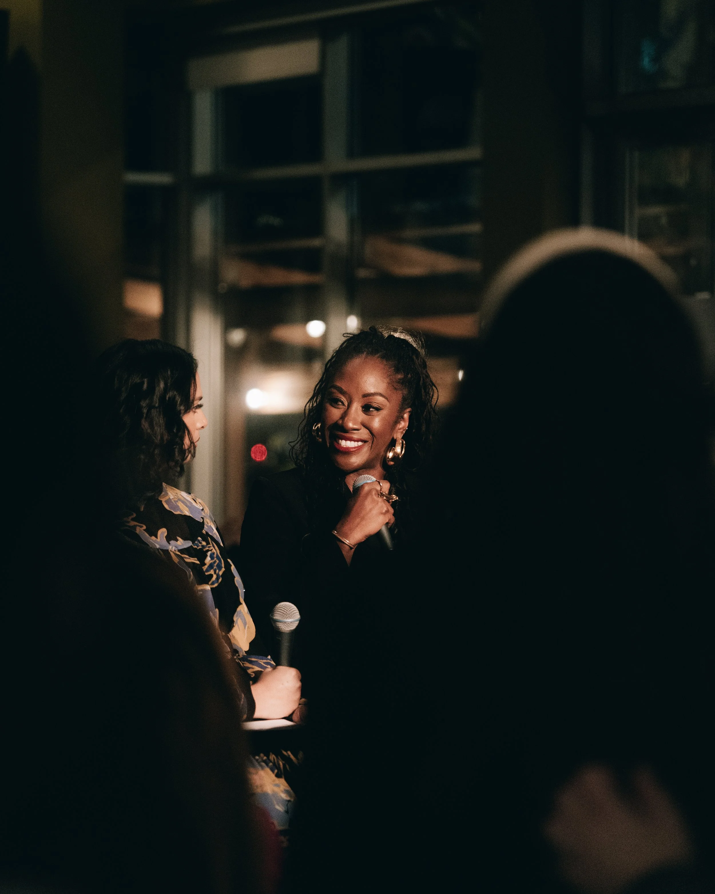 Two women having a conversation on stage at a microphone, smiling, in a dimly lit indoor setting.