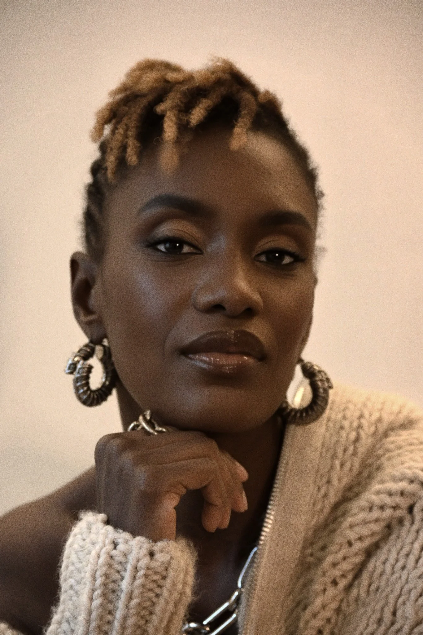 Close-up portrait of a woman with short, dreadlocked hair, wearing large hoop earrings, a beige knitted sweater, and a necklace, posing thoughtfully with her hand near her chin.